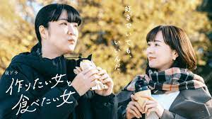 Two woman stand outside, holding sandwiches. in front of a background of gingko trees.