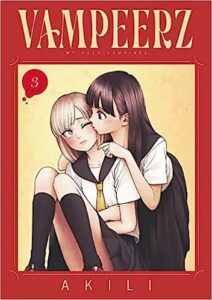 On a plain yellow background surrounded by a bright red frame, are two girls in white school sailor-style blouses and black school uniform skirts. One girl with long, dark hair, kisses the blonde girl on the upper cheek from behind.