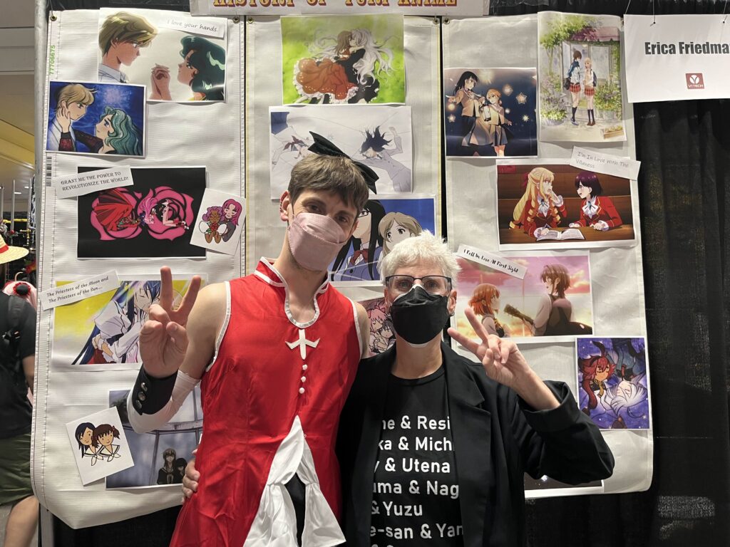 Matias Mintz as a magical girl and Erica Friedman posing in front of a wall of images from Yuri anime at Anime NYC 2024. Photo by Matias Mintz