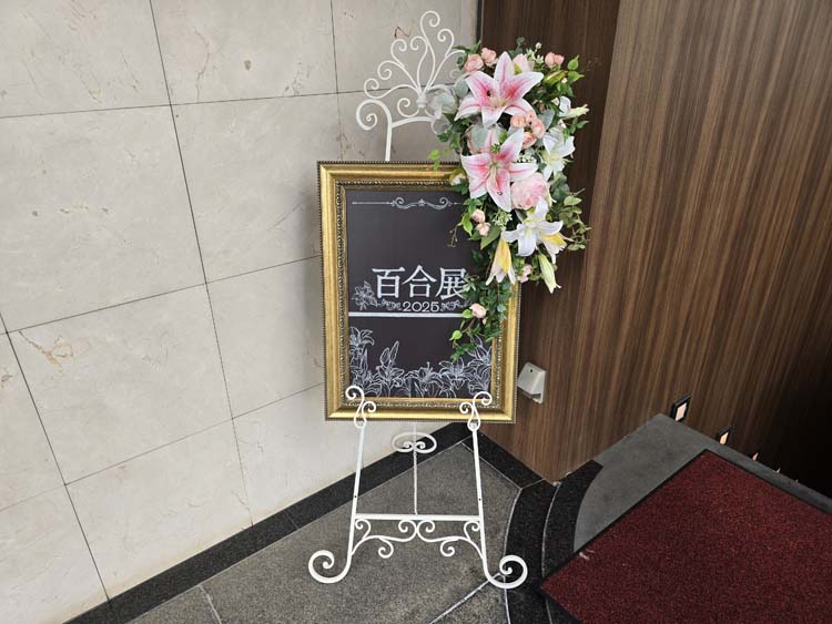 Wire stand with chalkboard sign reading Yuriten in Japanese , decorated with lilies. Yuriten Osaka 20253, B. Baker.