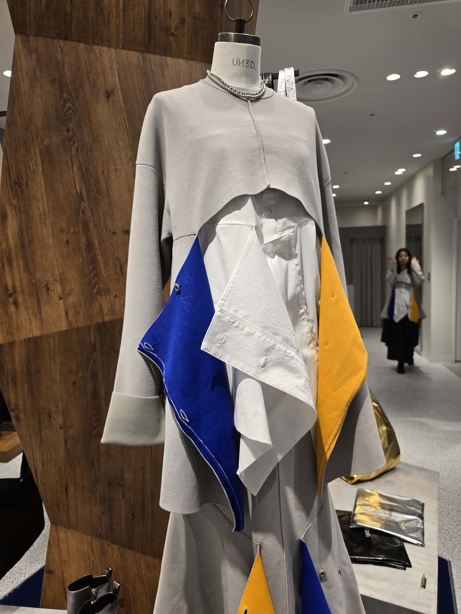 Artsy dress with hangining blue and yellow material under a white overpiece. Yuriten Osaka 2025, B. Baker.