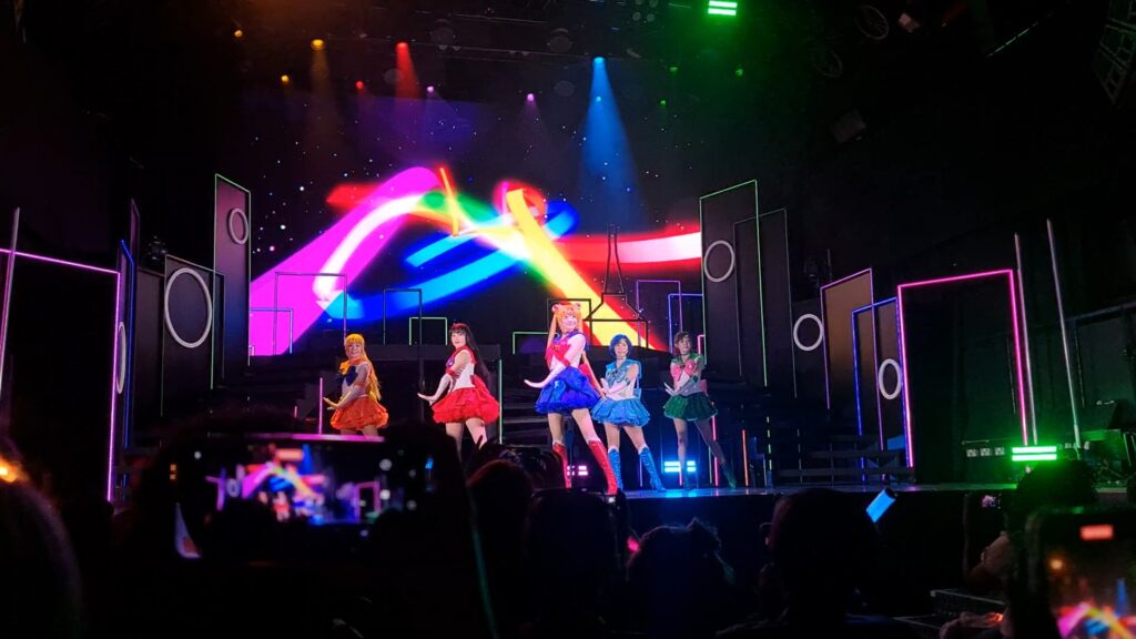 Sailor Moon Super Live, London 2025, photo by Ashley Payne. Five girls in Japanese sailor-suit school uniform inspired hero costumes, on a stage. Huge colorful ribbons of light fill the background.