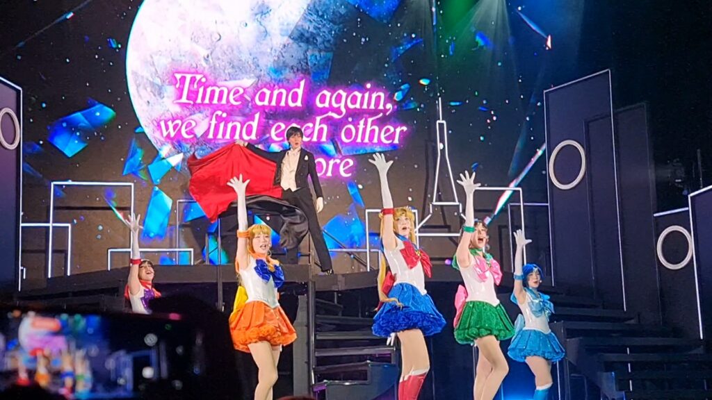 Sailor Moon Super Live, London 2025, photo by Rue Ball. Five girls in Japanese sailor-suit school uniform inspired hero costumes, on a stage, each posing in the traditional "appearance" pose of their character. Behind and above them is a person in a tuxedo and cape, while a large moon is projected behind them, superimposed with the words, Time and Again we find each other.