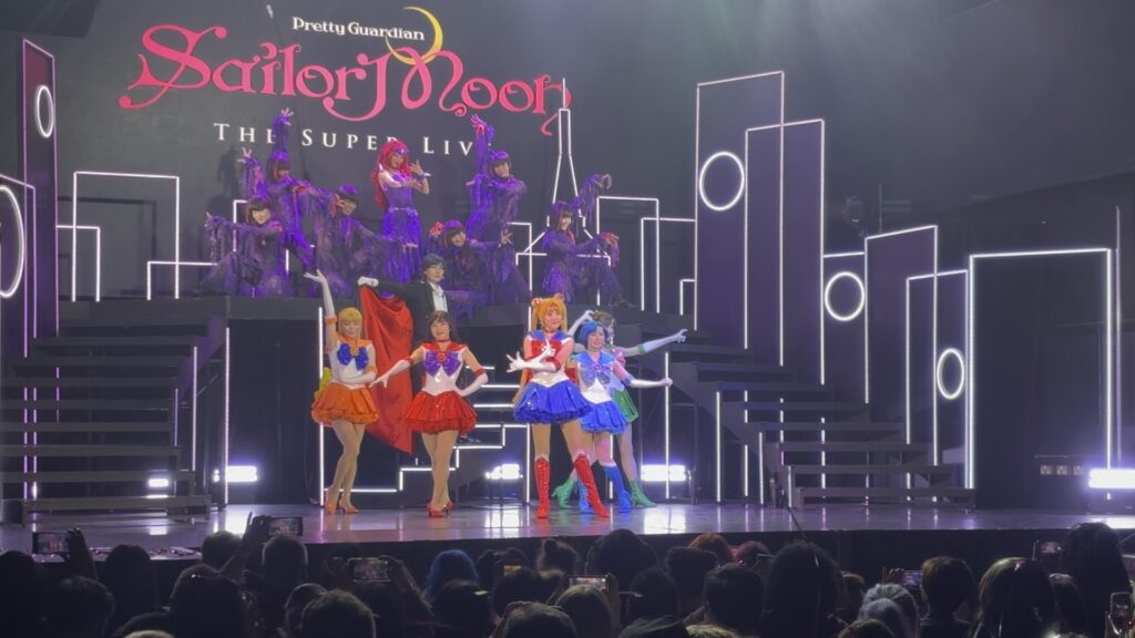 Sailor Moon Super Live, London 2025, photo by Rue Ball. Five girls in Japanese sailor-suit school uniform inspired hero costumes, on a stage, each posing in the traditional "appearance" pose of their character. Behind and above them are a woman in purple with scarlet hair and half a dozen people in purple posing around her.