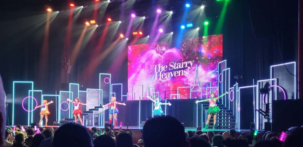 View of the stage at Sailor Moon The Super Live, 20205 at NJPAC in Newark, NJ. Five women in brightly colored super-hero version of girls "sailor style" Japanese school uniforms, sing and dance with a stage lit tot imply Tokyo, while a screen shows a deep-space image and the words "The Starry Heavens" behind them. Photo by P. Lawler, 2025.