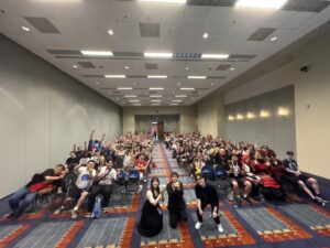 Photo of Miyuri Shimabukuro, Shinya Watada, and Hidetake Nishigaya posing in front of the crowd from the second RockLady panel at Otakon 2025. Shimabukuro-san and Watada-san are holding chibi dolls of the main cast.