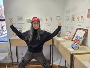 Rica Takashima at het Labo Brooklyn, standing spreadeagled surrounded by art for her solo exhibit for the 30th anniversary of "Rica 'tte Kanji!?", the first Yuri manga published in English.