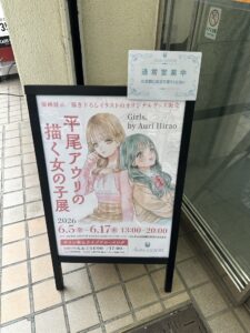 A signboard for Yuri Cafe Atelier Lililium, showing art by Hirao Auri for the inaugural art exhibit.