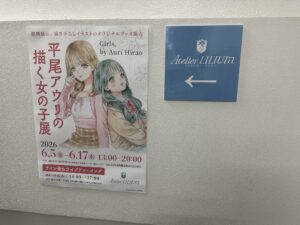 A hallway wall, with a poster for the Hirao Auri exhibit and a business sign in a cadet blue with whit letters that read Atelier Lilium, and a shield with a lily in white.