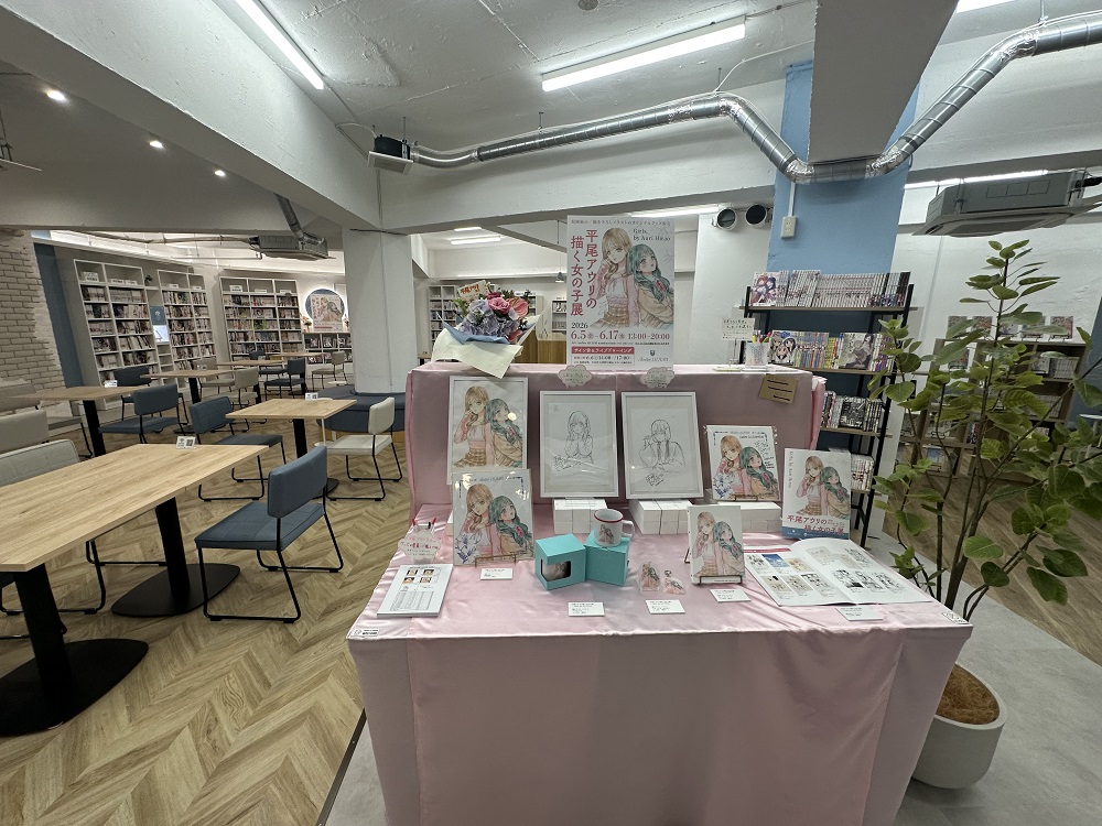 A view of the cafe and book space for Atelier Lililum. In the front is a table with a pink cloth featuring work and goods by Hirao Auri. Wooden tables on the left, feature gray and white chairs. Bookshelves along the wall are filled with books, in a well-lit white-wallled and ceilinged space.