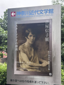 Poster of Yoshiya Nobuko exhibit at the Kanagawa Museum of Literature. Seated Yoshiya in a dressy blouse and fashionable short hair, looks at the camera with a slight smile. 