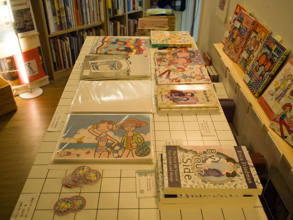 Table with Japanese and English language Yuri art and books, and pins with art by Rica Takashima.