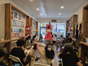 A gallery, filled with people doing figure drawing of two models dresses as characters from Touhou Project, Reimu in a red witchy maid costume and Marisa in black.